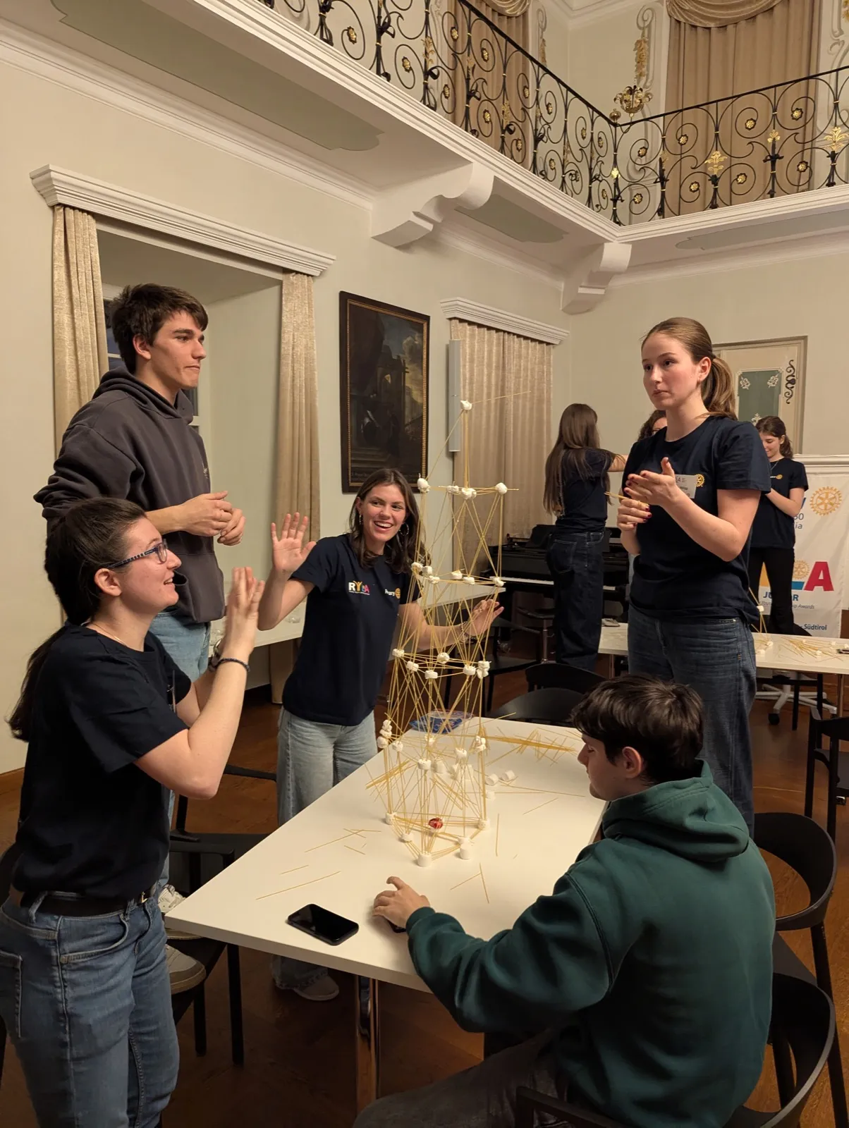 Students around a table in the monastery hall, raising their hands beside a tower of dry spaghetti and marshmallows that is still standing