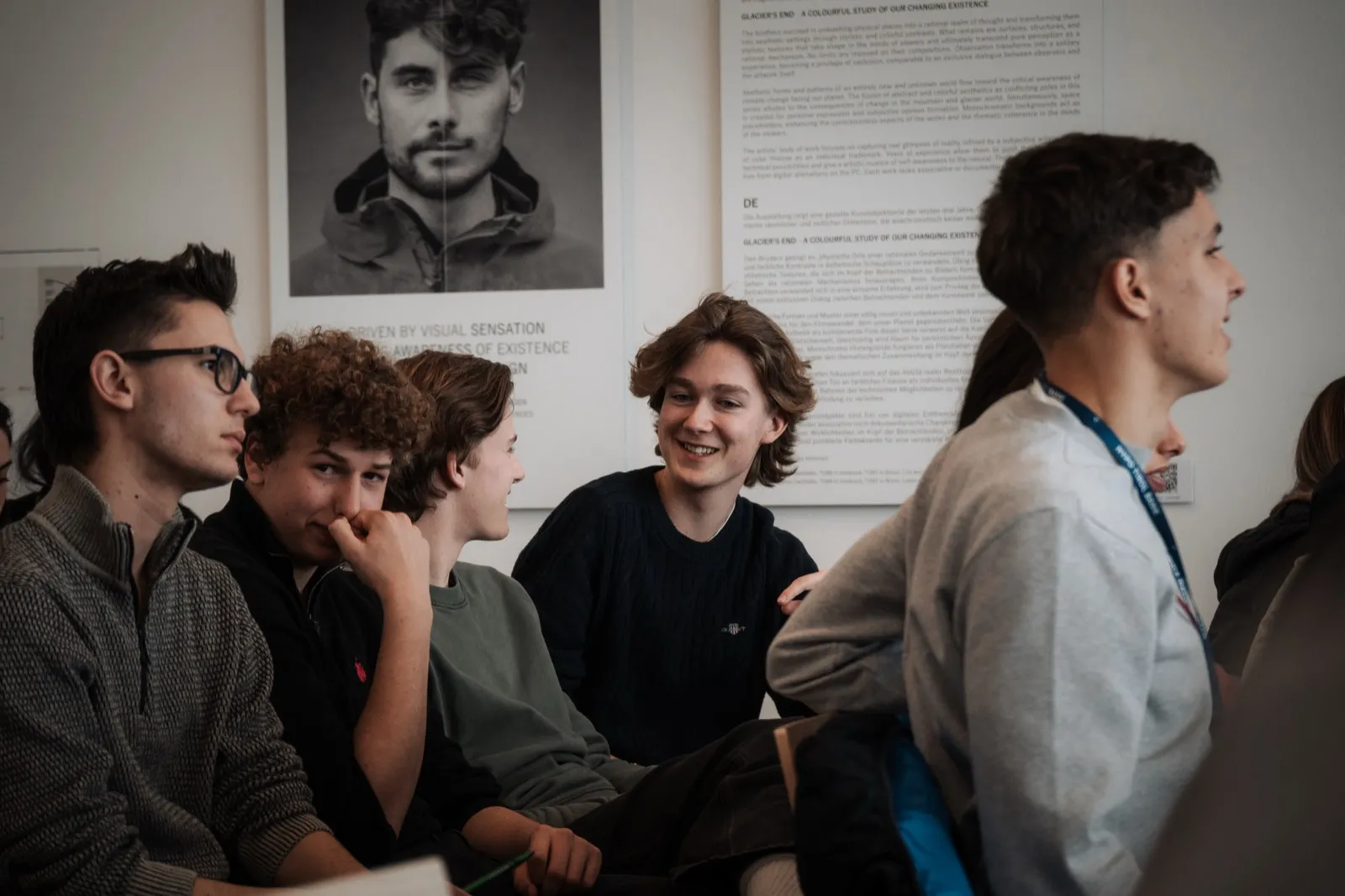 Participants sitting close together and listening, one of them smiling, in front of a wall carrying a large portrait print and a panel of text