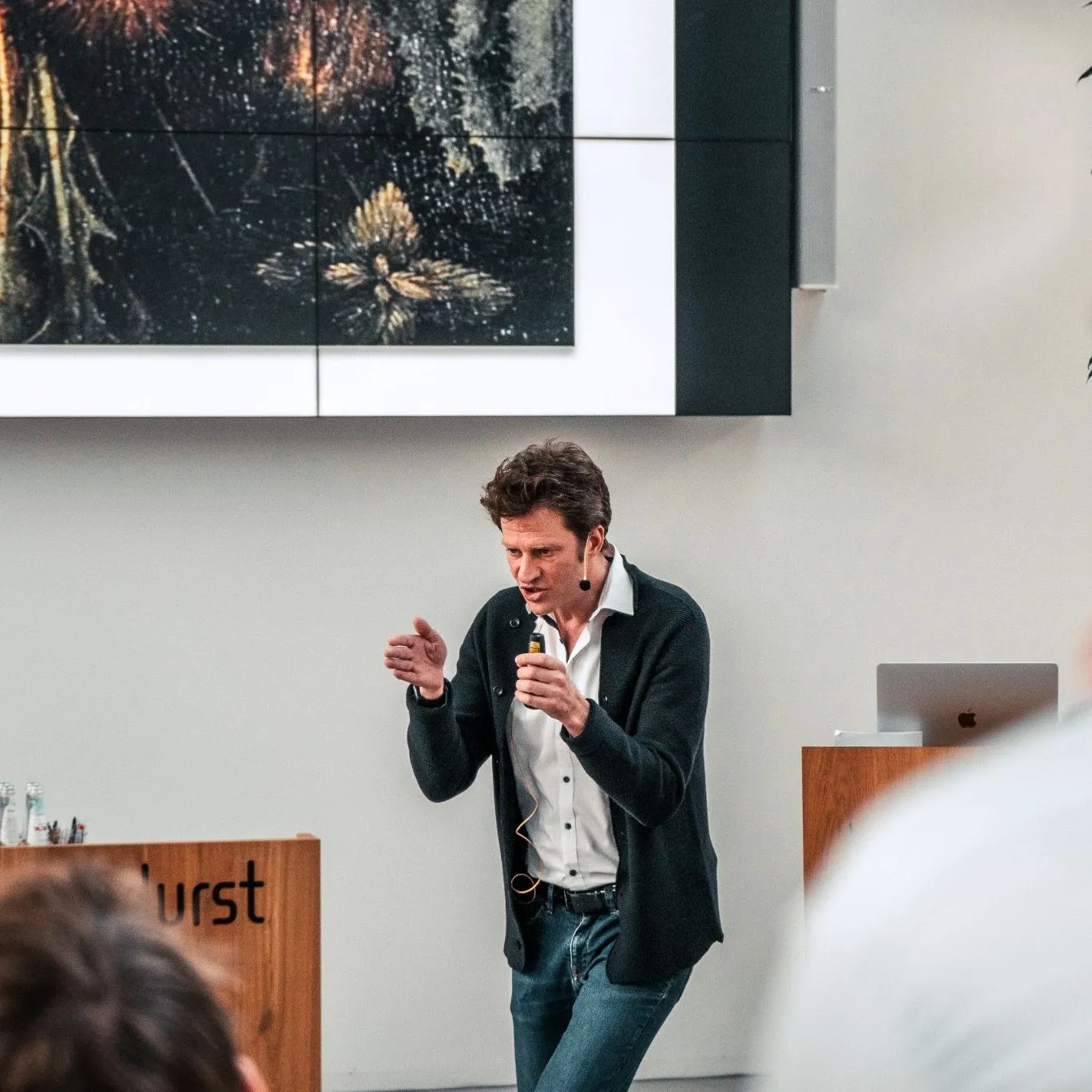 A speaker with a headset microphone talking and gesturing in front of a large photographic print, beside a lectern carrying the Durst name, with heads of the audience in the foreground