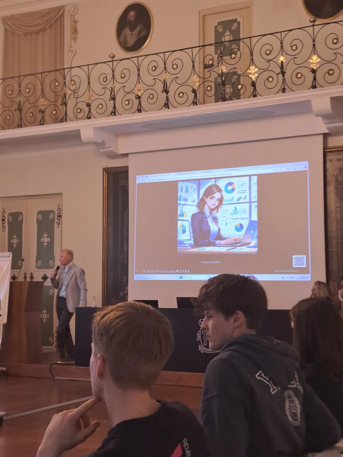 A speaker with a microphone beside a projected slide, seen over the heads of participants in the front row