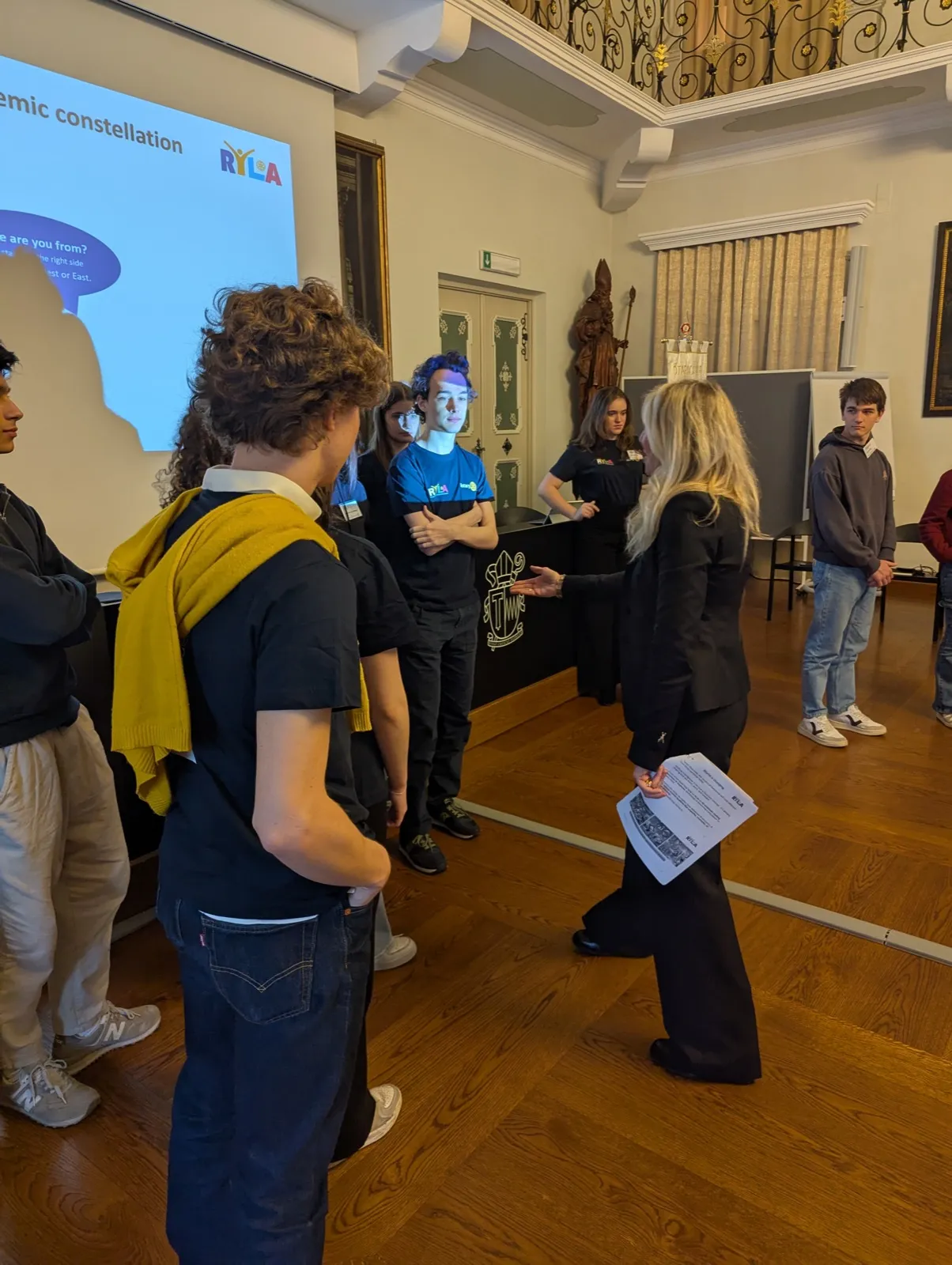 Participants standing in a loose circle in a hall while a woman holding a sheet of paper speaks to them, a projected slide with the RYLA logo behind